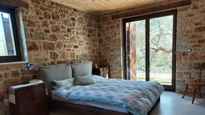 A bedroom with a stone wall, a bed with a patterned bedspread, a wooden dresser, and a window with a view of trees. - Isolated luxury villa with pool, 30 mins to sea, 2 km to village shops tavernas (messenia)