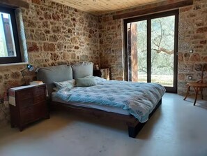 Een slaapkamer met een stenen muur, een bed met een beddeken met een patroon, een houten dressoir en een raam met uitzicht op bomen.