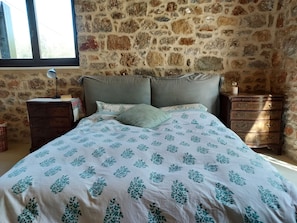 A bedroom with a stone wall, a bed with a patterned bedspread, a bedside lamp, and a dresser.