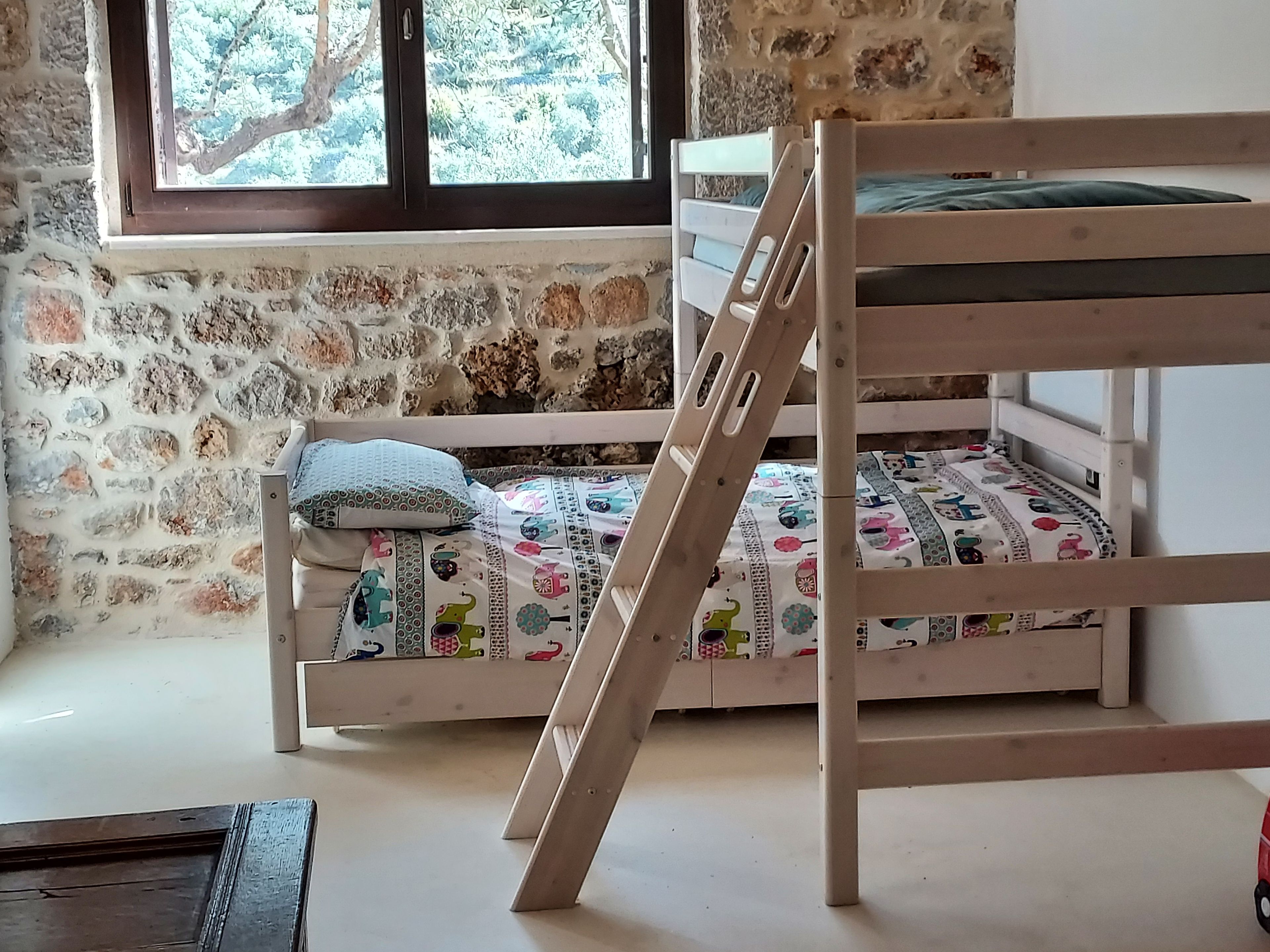 Uma cama de beliche com escada de madeira, janela com vista para árvores e parede de pedra.
