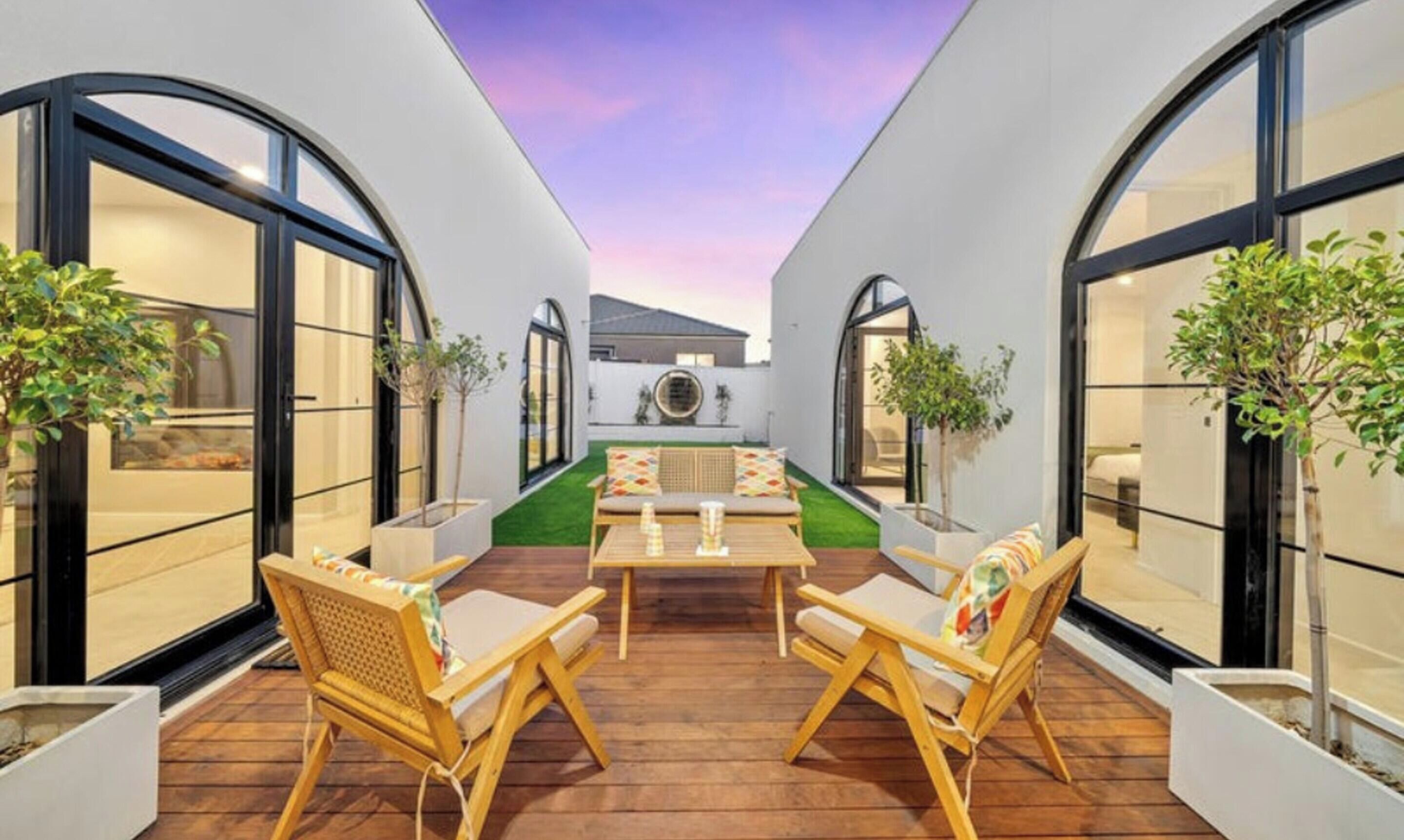 A modern outdoor patio with a wooden table and chairs, surrounded by white walls and large windows.