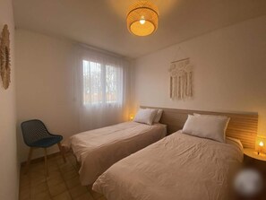 A room with two beds, a chair, a window with curtains, a wall hanging, and a hanging light fixture. - L'ESTIMADA - 4 CHAMBRES - PISCINE (Argelès-sur-Mer)