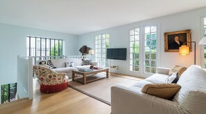 Ein geräumiges Wohnzimmer mit einer Couch, einem Sessel und einem Couchtisch, großen Fenstern und einem Gemälde an der Wand.