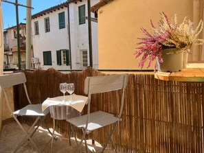 Ein Balkon mit zwei Stühlen und einem Tisch, ein Blumentopf mit rosa Blüten und ein Gebäude im Hintergrund.