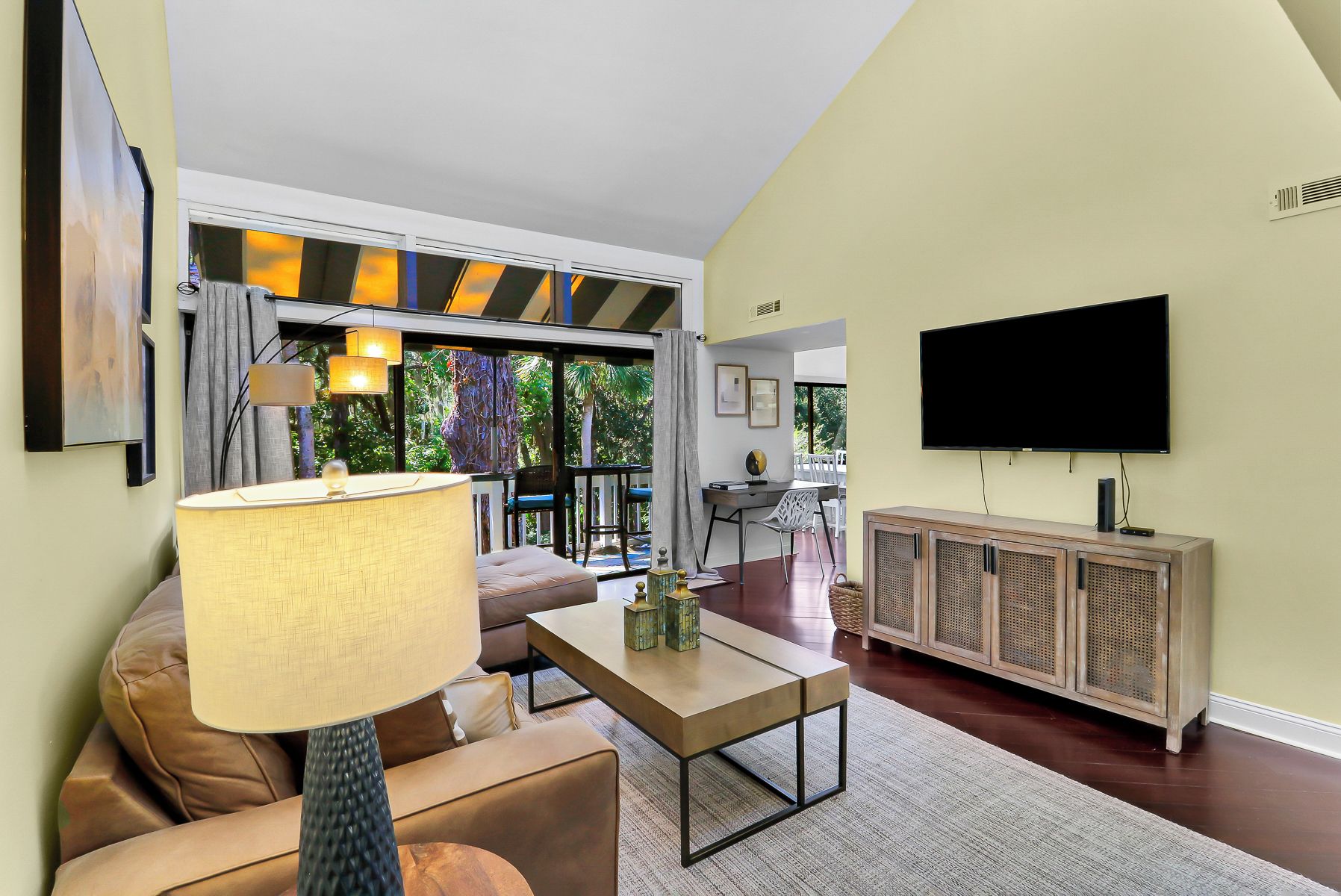 A modern living room with a sofa, coffee table, and a television. There is a dining area with chairs and a table visible through a glass door.