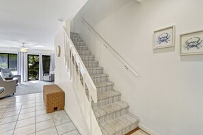 Eine Treppe mit weißer Geländer führt zu einer oberen Etage, am Fuß befindet sich eine gewebte Sitzbank und an der Wand hängen zwei gerahmte Kunstwerke.