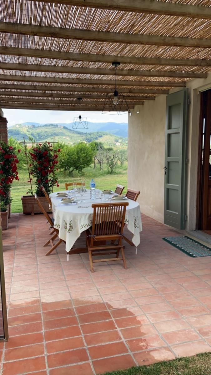 Um pátio externo com mesa posta para refeições, rodeado de cadeiras e vista para o campo.