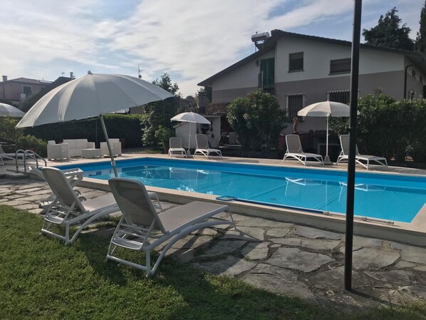 Una piscina con sillas de descanso y sombrillas, rodeada de pavimento de piedra y follaje.