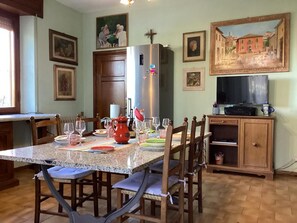 A dining area with a granite table, wooden chairs, a refrigerator, and a television on a wooden cabinet. - Casa Riccardo in Villa (BARGA)