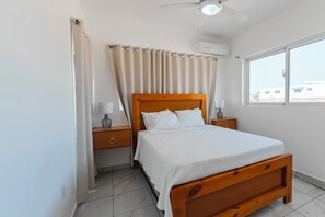 Ein Schlafzimmer mit einem hölzernen Bett, zwei Nachttischen mit Lampen, einem Deckenventilator, einem Fenster mit Aussicht und einem Vorhang.