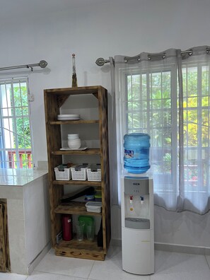 A room with a wooden shelf unit, a water dispenser, and a window with a view of greenery.