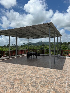 Un espace extérieur abrité, doté d’un sol carrelé, d’une table avec des chaises et offrant une vue sur des collines verdoyantes en arrière-plan.