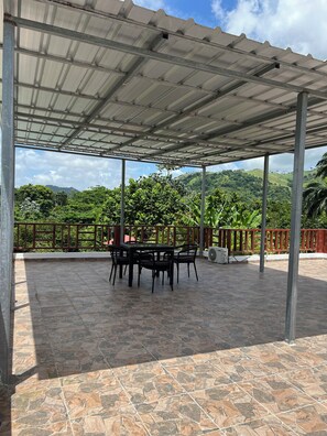 Un patio exterior techado con una mesa y sillas, rodeado por una barandilla metálica y vistas a un paisaje montañoso.