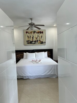 A hotel room with a bed, white linens, a ceiling fan, and a wall-mounted artwork.
