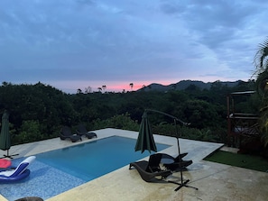 Una piscina con sillones de descanso y sombrillas, rodeada de abundante vegetación y colinas al fondo.