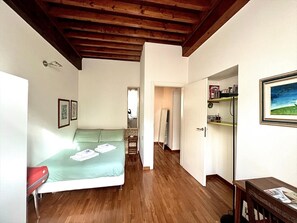 Ein Zimmer mit einem Bett, Holzboden, einer Tür zu einem anderen Raum und einem Regal mit Gegenständen.