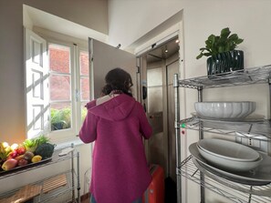 Uma pessoa em uma cozinha com janela, geladeira e prateleiras com louças.
