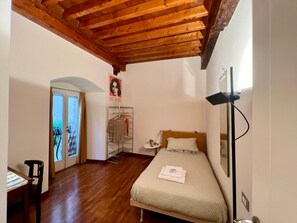 Quarto com teto de madeira, cama com colcha branca e bege estampada, mesa de cabeceira, abajur e um pôster na parede.