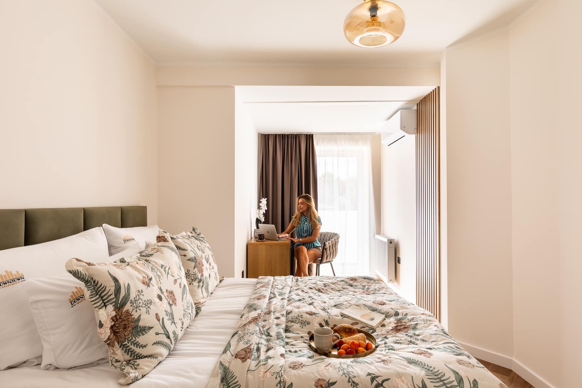 A hotel room with a bed, a desk, and a person working on a laptop.
