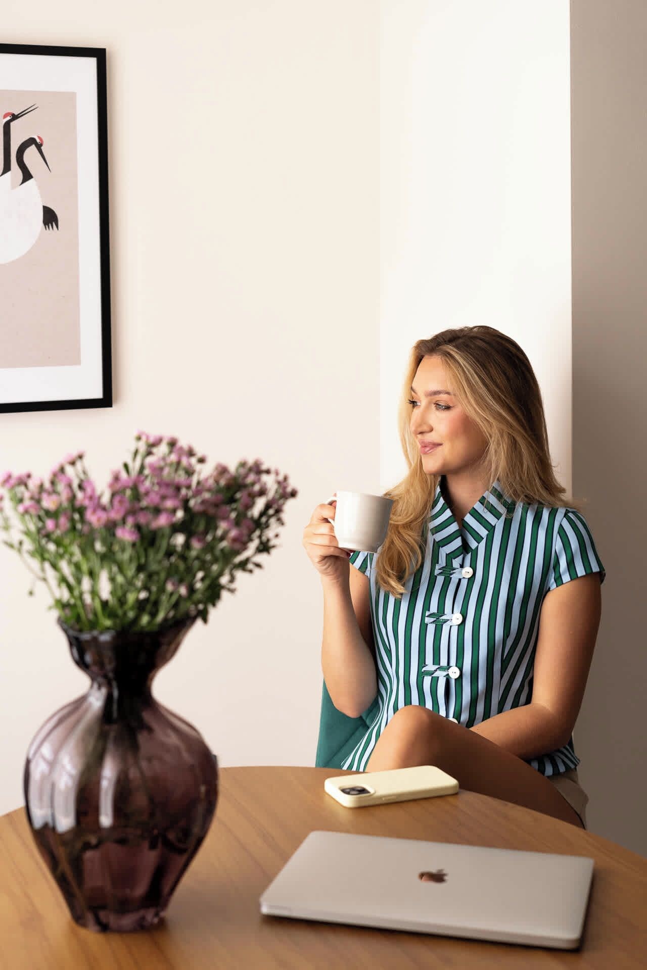 A person sitting at a table with a laptop, holding a mug, and a vase with flowers.