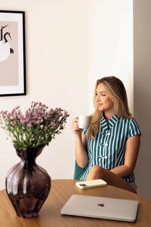 En person sidder ved et bord med en bærbar computer, holder en kop, og der står en vase med blomster.