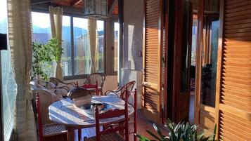 A sunlit patio with a round table and chairs, a potted plant, and wooden shutters.