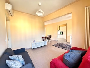 Ein Wohnzimmer mit einer Couch, einem roten Sessel, einem Essbereich mit Stühlen und einem Tisch sowie einer Küche im Hintergrund.