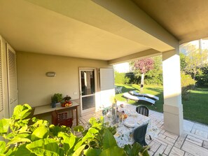 Une terrasse couverte avec une table dressée pour les repas, une porte donnant sur l’extérieur et une vue sur le jardin.