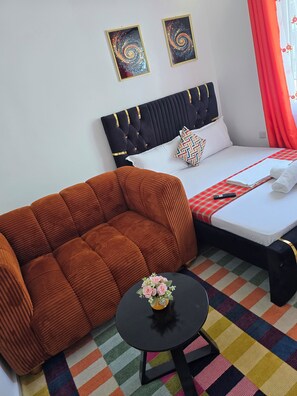 A hotel room with a bed, a sofa, a small table with a vase, and two wall art pieces. - Matunda homes 
Cozy.Classy.Beautiful (Mombasa)