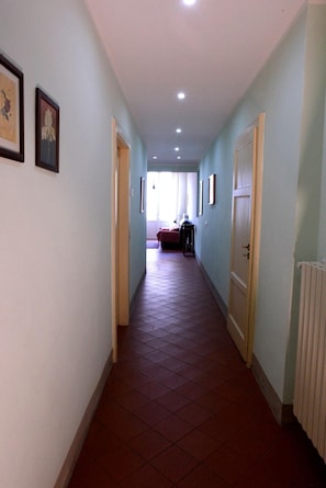 Ein Korridor mit Türen an beiden Seiten, einem Heizkörper rechts und einem Bett mit roter Decke in der Ferne.