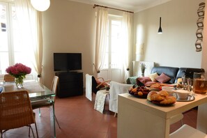 Een woonkamer met een bank, een televisie, een eettafel met stoelen, en een klein keukentje met een ontbijt gedekte tafel.