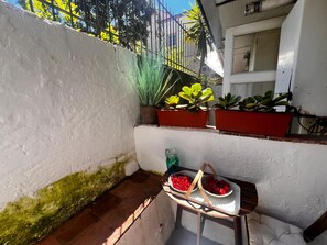 Un petit espace patio avec un mur blanc, une table en bois sur laquelle se trouve un panier rempli de fruits rouges, et plusieurs plantes en pot.