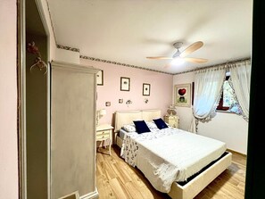 Een slaapkamer met een bed, nachtkastje, lamp, plafondventilator en een raam met gordijnen.