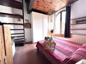 Une pièce comprenant un lit, un banc en bois, une petite table avec quelques objets, une fenêtre avec des rideaux et une étagère fixée au mur.