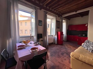 A dining area with a table set for two, a kitchen with red cabinets, and a sofa in the living area. - civico 12 appartamento in centro storico (LUCCA)