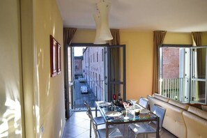 A dining area with a glass table and chairs, a sofa, and a view of a narrow street through an open door. - Casa ai giardini in front of the ancient wall (LUCCA)
