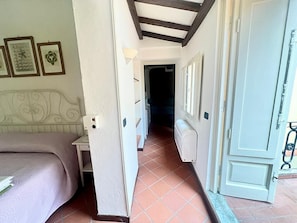 Quarto de hotel com cama, radiador, porta e corredor que leva a outro quarto.