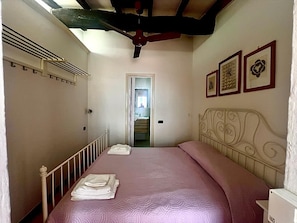 Quarto com cama, guarda-roupa, ventoinha no teto e porta que dá para uma escadaria.