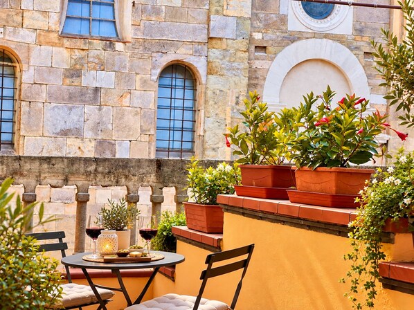 Edifício de pedra com janelas em arco e uma varanda com plantas em vasos e uma mesa com cadeiras.