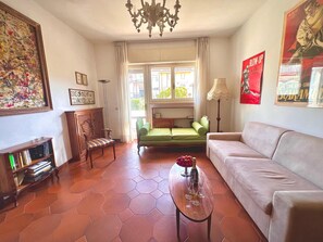 Una sala de estar con un sofá, un sofá verde, una silla de madera, una mesa de centro con una botella y vasos, una estantería y un cuadro grande.