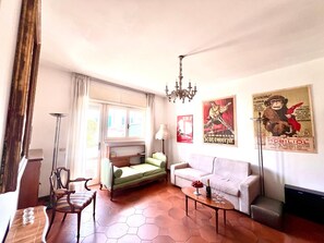 Un salon comprenant un canapé, un fauteuil, une table basse, une lampe et un lustre. Les murs sont ornés de posters et il y a une fenêtre avec des rideaux.