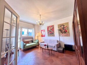 A living room with a sofa, a green armchair, a coffee table, and a chandelier. There are posters on the wall and a window with curtains. - Casa Sant'Andrea nel cuore di Viareggio (VIAREGGIO)