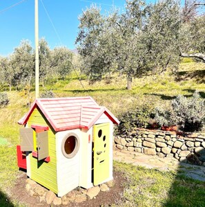 A small, colorful birdhouse with a red roof and a circular entrance, situated in a garden with a stone wall and olive trees in the background. - Casa Pozzi in the heart of the Lucca hills (Capannori)