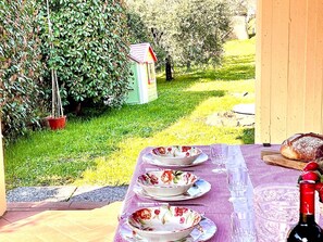 Un espace extérieur aménagé pour prendre les repas, avec une table dressée pour deux personnes, entouré d’un jardin agrémenté d’un petit abri de jardin et d’une plante en pot.