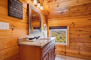 A wooden cabin bathroom with a granite countertop, a large mirror, and a view of a forest through a window. - New Listing, Hot Tub, Private Indoor Heated Pool, Game Room, Theater Room (Pigeon Forge)