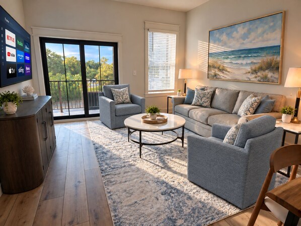 Un salon comprenant un canapé, des fauteuils, une table basse, un téléviseur et un tableau représentant une scène de plage.