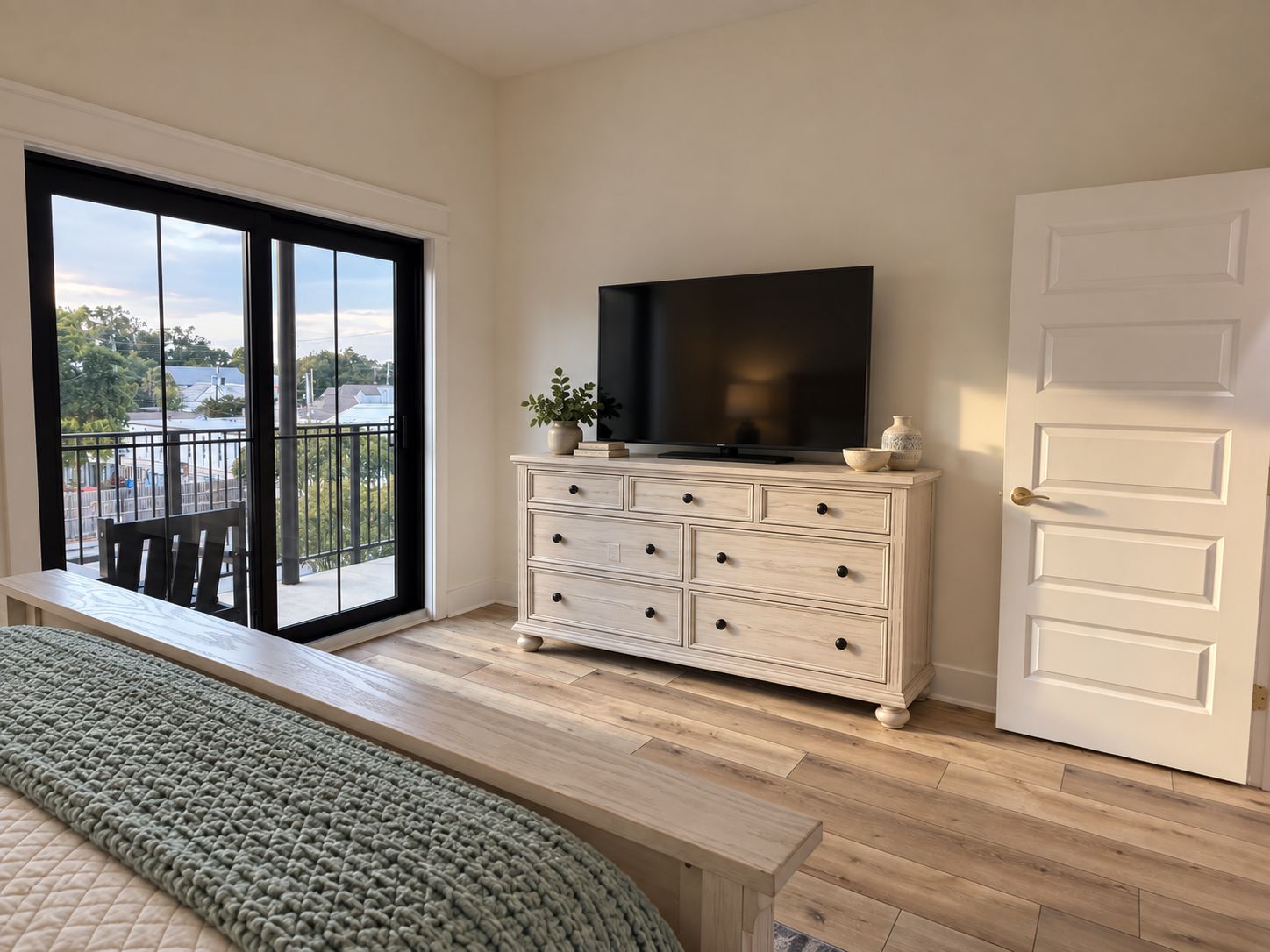 A room with a large sliding glass door leading to a balcony, a white dresser with a flat-screen TV, a bench, and a door.