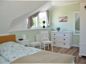 A bedroom with a sloped ceiling, a bed with floral bedding, a white dresser, a chair, a nightstand, and a window with curtains. - Holiday Home in Zingst With Sauna (Zingst)