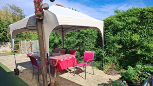 Un espace patio extérieur avec une table et des chaises sous une tonnelle blanche, entouré de verdure et de plantes en pot.