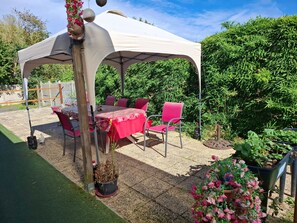 Un espace patio extérieur avec une table et des chaises sous une tonnelle blanche, entouré de verdure et de plantes en pot.